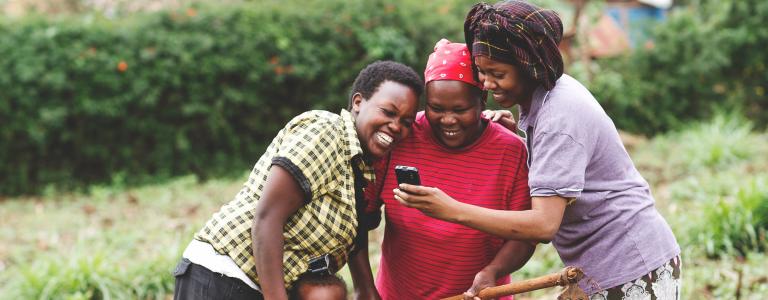 Three women on a farm in Kenya look at a mobile phone while smiling as a small child stands beneath