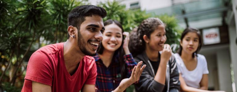 Four young adults sit on a bench outside talking and laughing