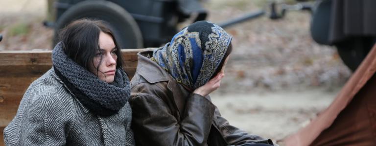 Two women sit side-by-side in grey clothing, one in a headscarf is turned away and covering her mouth with her hand