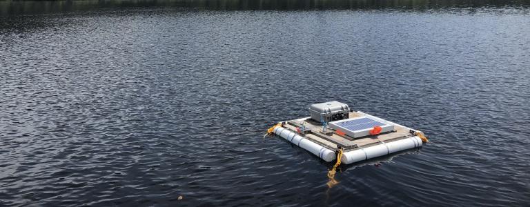 Floating platform with a solar panel sits on a lake in front of trees and a cloudy sky