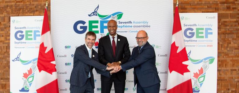 Three men in suits awkwardly hold hands in front of two Canadian flags