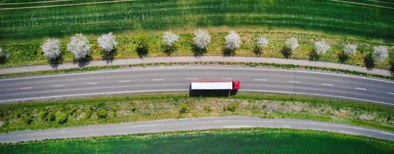 A semi-truck drives along a road surrounded by green landscape.