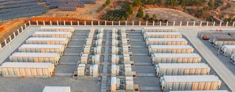 An energy storage facility stands in front of a field of solar panels.